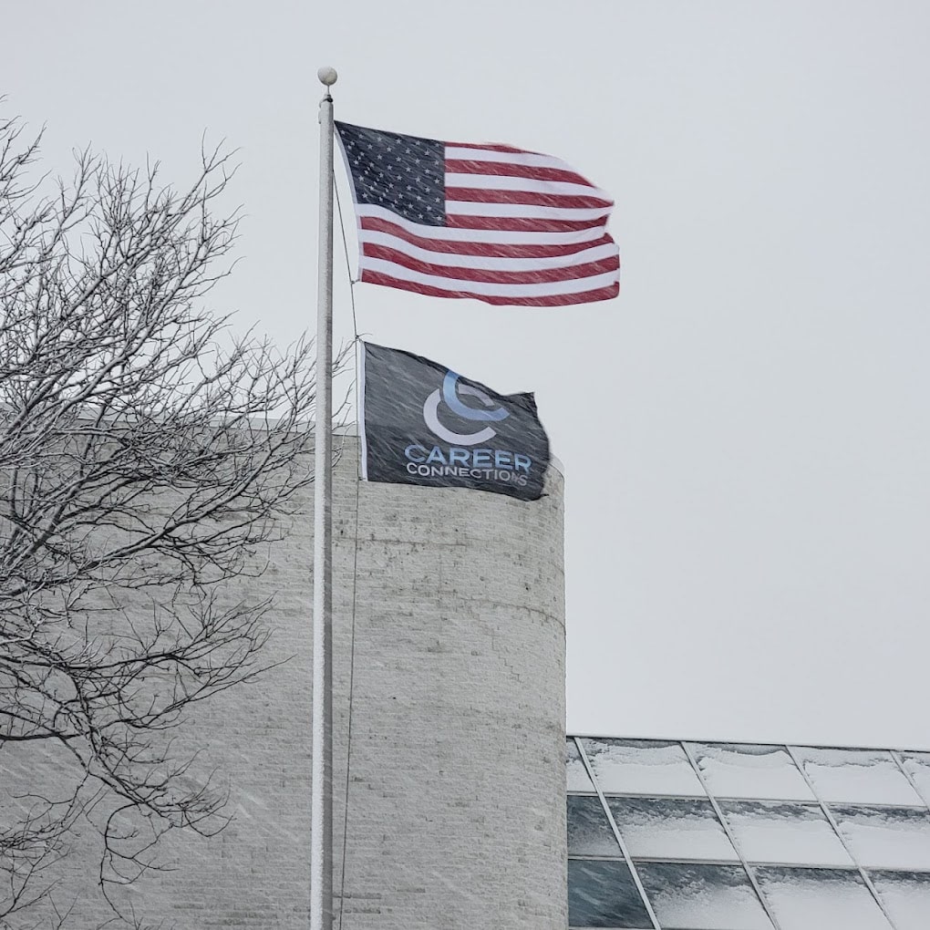 Two flags on a tall flagpole outside the Career Connections office building on a snowy day - one American Flag and the other with the Career Connections Staffing Services logo on a black background