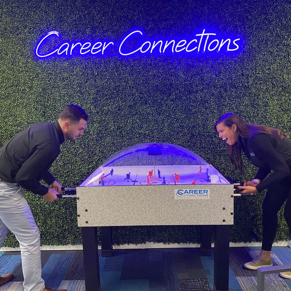 Two Career Connections employees, one male and one female, play a lively game at a dome hockey table in the Career Connections office with a 'Career Connections' neon sign in the background.
