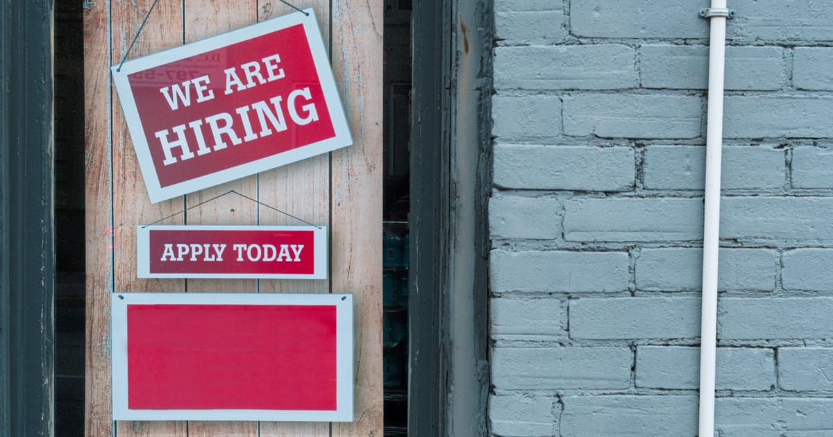 Three red signs on a weathered wood door, two saying "We Are Hiring" and "Apply Today", next to a gray brick wall
