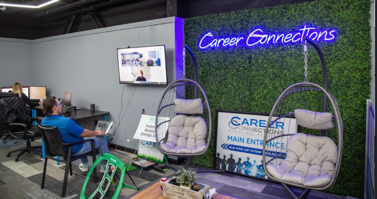 Modern career center with a green artificial hedge wall featuring a neon "Career Connections" sign. Two hanging cushioned chairs sit in front, creating a casual lounge area. To the left, two people work at desks with a wall-mounted screen showing a virtual meeting. Directional signs and decor elements like "Professional Storage Area" and "Our Happy Place" add personality to the well-lit, contemporary space.