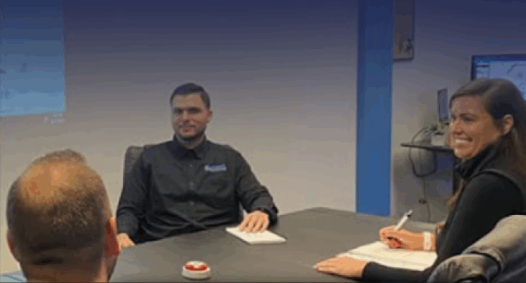 Four individuals seated around a conference table in a professional office setting. One person, wearing a black shirt with a logo, faces the camera and appears to lead a discussion. Another writes in a notebook, while a third is partially visible with their back to the camera. A red button sits on the table, and a computer monitor and office equipment are visible in the background, suggesting a collaborative meeting or brainstorming session.